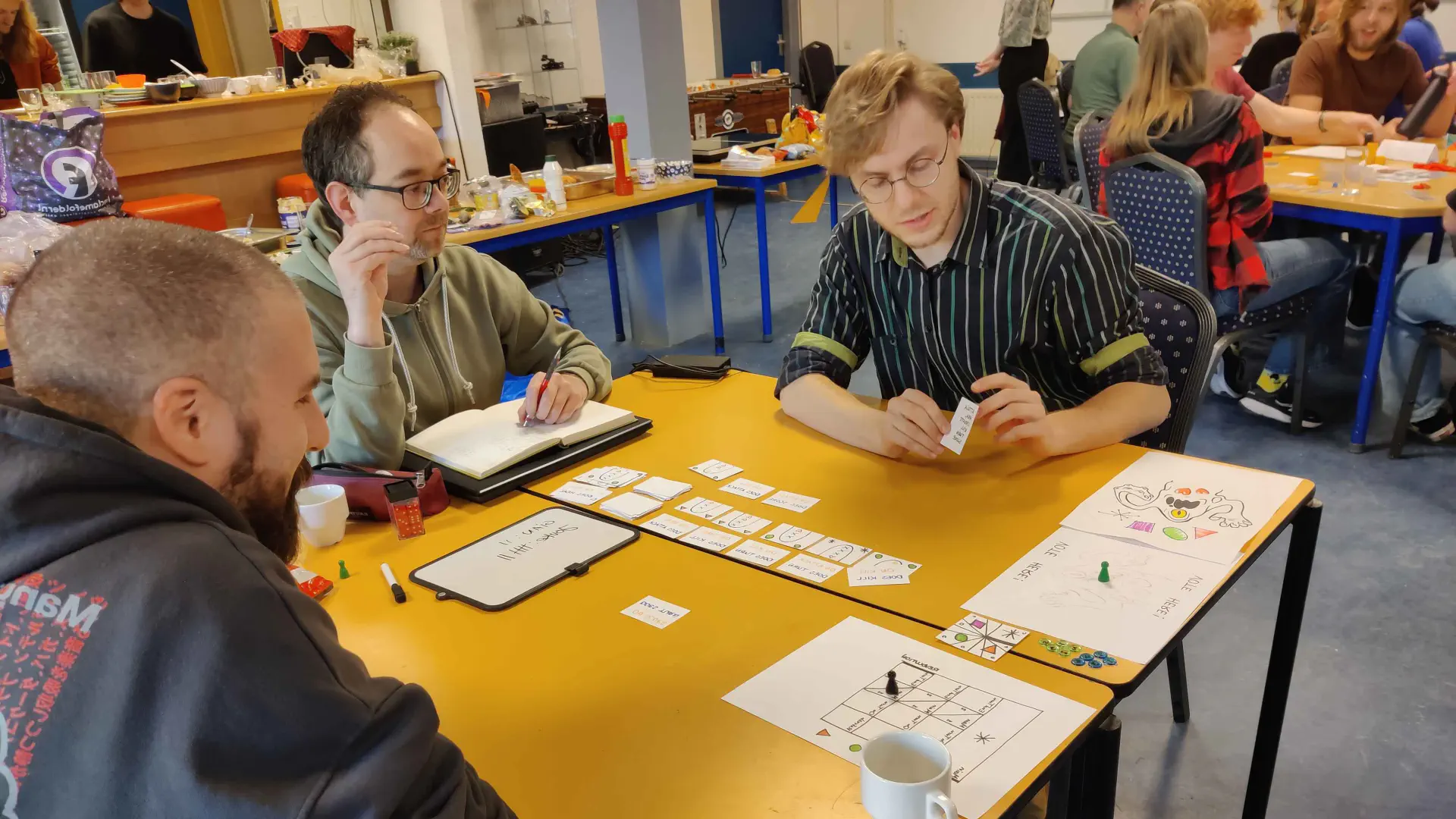 Three people sitting at a table testing a selfmade board game. One of them is holding a small paper, probably part of the game.